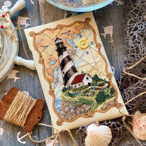 May include: Cross-stitch art featuring a lighthouse with black and white stripes, a red-roofed house, and a yellow compass rose. The artwork is framed with a rope design and set against a map background. The scene includes birds and is surrounded by nautical elements.