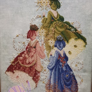 May include: Three embroidered figures in gowns, each holding a star-shaped object, stand on a white background with a light blue tint. The figures are wearing green, pink, and blue gowns with gold accents. The background is covered in small white stars.