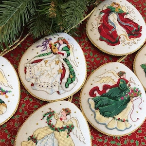May include: Six round, white, embroidered ornaments with gold trim, each featuring a different angel design. The angels are dressed in various colours, including red, green, and white, and are holding objects such as musical instruments and flowers.