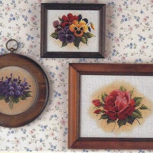 May include: Three framed cross-stitch embroidery designs featuring floral bouquets. The top frame shows a bouquet of yellow and purple pansies. The middle frame shows a bouquet of purple violets. The bottom frame shows a single red rose.