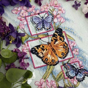 Può includere: Ricamo a punto croce con tre farfalle in cornici quadrate, con ali arancioni, viola e blu. Lo sfondo è un tessuto azzurro con fiori rosa e fogliame viola.