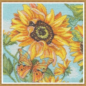 Puede incluir: Punto de cruz de girasoles y una mariposa. Los girasoles son amarillos y naranjas con centros marrones, sobre un fondo azul claro. Una mariposa naranja y azul está colocada cerca de las flores. La obra está enmarcada.