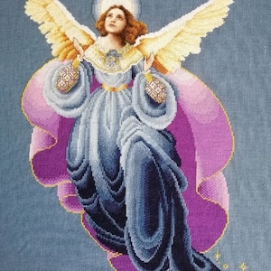 May include: A cross-stitch embroidery depicting an angel with outstretched wings, wearing a blue and purple gown with a gold belt and halo. The angel is surrounded by a pink and purple background with gold stars.