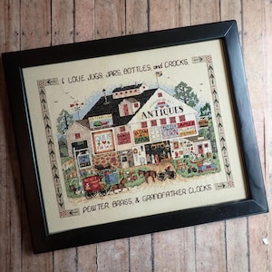 May include: Framed cross-stitch artwork showing an antique shop with the word "ANTIQUES" on the building. The artwork includes text that reads "I LOVE JUGS, JARS, BOTTLES, and CROCKS" and "PEWTER, BRASS, & GRANDFATHER CLOCKS". The shop has signs for quilts, grain, corn, and other items.