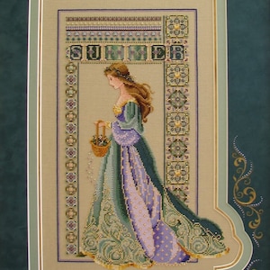 May include: Cross-stitch embroidery of a woman in a green and purple dress holding a basket of flowers. The embroidery is framed and the text "SUMMER" is stitched above the woman.