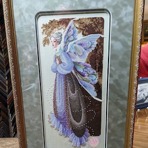 Può includere: Un ricamo a punto croce incorniciato che rappresenta una fata con ali iridescenti e un vestito viola. La fata tiene un libro ed è circondata da uno sfondo di foresta marrone e verde. La cornice è dorata con un bordo decorativo.