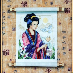 May include: Cross-stitch artwork of an Asian woman in a colorful robe, holding a dove. The artwork is framed and mounted on a scroll with wooden dowels. The background features Chinese characters and a textured pattern.