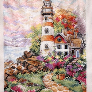 May include: Cross-stitch of a lighthouse with red and white stripes, a small house, and a stone path. The scene is surrounded by colorful flowers and greenery. The sky is a mix of pink, orange, and blue.