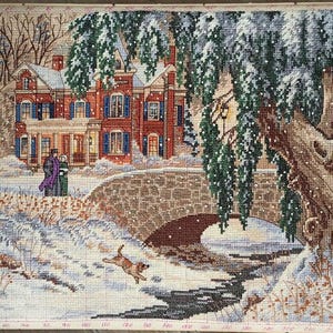 May include: A cross-stitch winter scene depicts a snow-covered landscape with a brick house, bridge, and a flowing stream. A couple walks on a path, and a rabbit jumps across the stream. The scene is framed by trees.