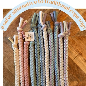 May include: A collection of braided dog toys in various pastel colors, including pink, blue, and green. Each toy has a knotted end and is made of soft, woven material. Text reads: "a safer alternative to traditional rope toys."