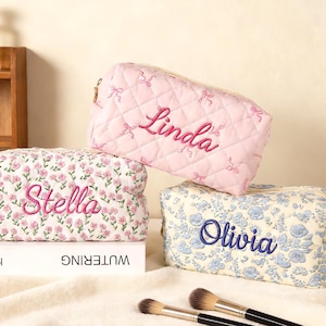 Könnte beinhalten: Drei personalisierte Kosmetiktaschen in Rosa, Weiß mit Blumenmuster und Blau mit Blumenmuster. Die rosa Tasche ist gesteppt und mit dem Namen "Linda" bestickt. Die weiße Blumentasche hat "Stella" und die blaue Blumentasche hat "Olivia". Make-up-Pinsel sind im Vordergrund.