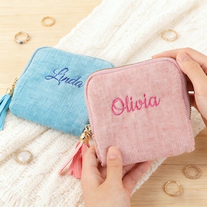 May include: Two small, square, zippered pouches. One is light blue with "Linda" embroidered in blue, the other is pink with "Olivia" embroidered in pink. Both have gold zipper pulls and matching tassels. Several rings are scattered around the pouches.