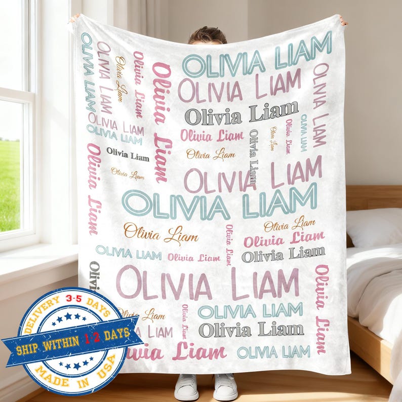 Op de afbeelding: Een witte deken met de namen "Olivia Liam" in verschillende kleuren en lettertypen. De namen worden herhaald over de deken. De deken wordt door een persoon omhoog gehouden. De deken heeft een leveringsbadge met de tekst "Verzending binnen 1-2 dagen".