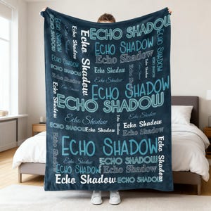 Op de afbeelding: Een donkerblauwe deken met de woorden "ECHO SHADOW" in verschillende maten en tinten blauw en wit. De deken wordt getoond in een slaapkamer, vastgehouden door een persoon.