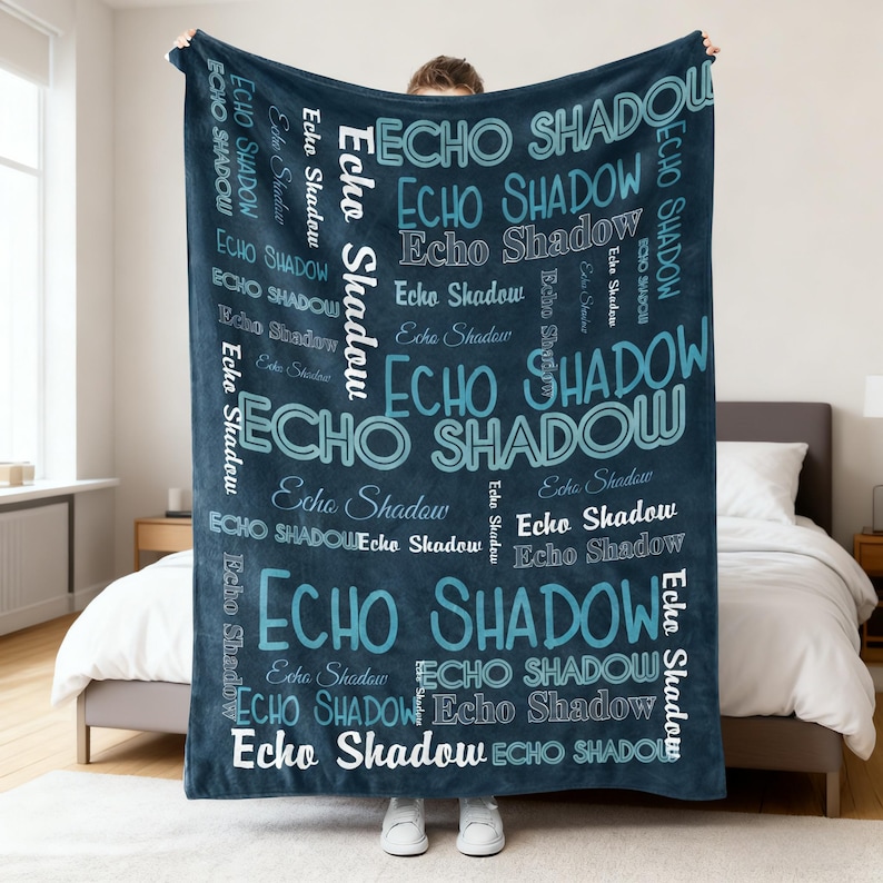 Op de afbeelding: Een donkerblauwe deken met de woorden "ECHO SHADOW" in verschillende maten en tinten blauw en wit. De deken wordt getoond in een slaapkamer, vastgehouden door een persoon.