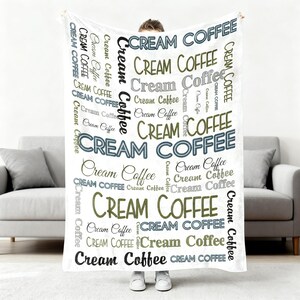Op de afbeelding: Een witte deken met de woorden "Cream Coffee" in verschillende lettertypen en kleuren, waaronder zwart, grijs en olijfgroen. De deken wordt vastgehouden door een persoon in witte schoenen, voor een grijze bank.