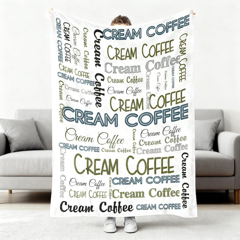 Op de afbeelding: Een witte deken met de woorden "Cream Coffee" in verschillende lettertypen en kleuren, waaronder zwart, grijs en olijfgroen. De deken wordt vastgehouden door een persoon in witte schoenen, voor een grijze bank.