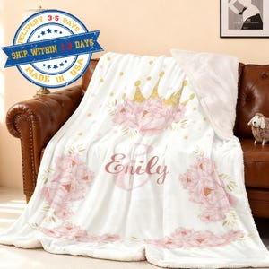 Könnte beinhalten: Weiche, weiße Decke mit rosa Blumenmuster, goldener Krone und dem Namen "Emily" in rosa Schrift. Die Decke liegt auf einem braunen Ledersofa. Hergestellt in den USA.