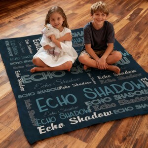 Op de afbeelding: Een marineblauwe deken met de woorden "Echo Shadow" in verschillende lettertypen. Twee kinderen zitten op de deken. E&eacute;n kind houdt een wit eenhoornknuffel vast.