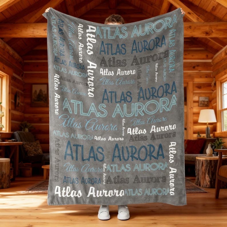 Op de afbeelding: Een grijze deken met de naam "ATLAS AURORA" in verschillende lettertypen en tinten blauw en wit. De deken wordt omhoog gehouden, met een houten hutinterieur op de achtergrond.