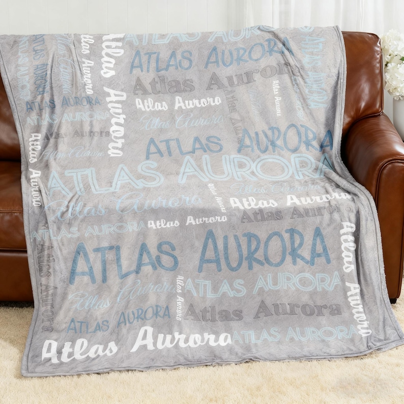 Op de afbeelding: Een grijze fleece deken met de namen "Atlas" en "Aurora" in verschillende lettertypen en grijstinten en blauw. De deken is over een bruine leren bank gedrapeerd, een gezellig en gepersonaliseerd woondecoratie-item.