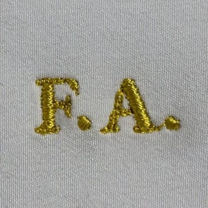 Op de afbeelding: Goud geborduurde initialen "F.A." op een witte stoffen achtergrond.