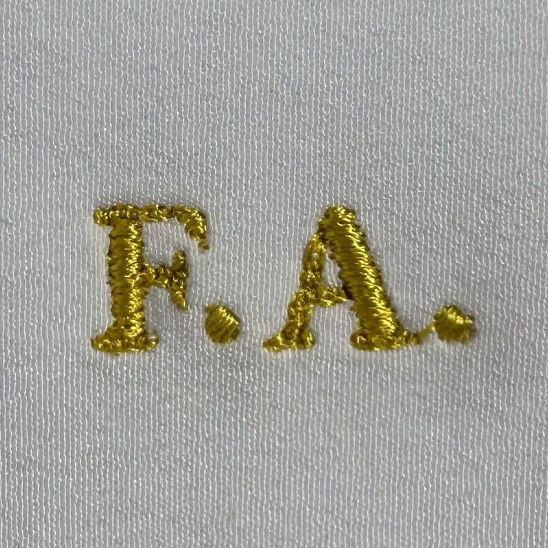 Op de afbeelding: Goud geborduurde initialen "F.A." op een witte stoffen achtergrond.