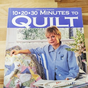 May include: A book titled "10-20-30 Minutes to Quilt" by Nancy Zieman. The cover features a woman in a blue shirt, sitting on a white wicker chair with a colorful quilt. The book is on a wooden surface.