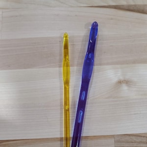 May include: Two crochet hooks, one yellow and one blue, are displayed on a light wood surface. The hooks are made of transparent plastic and have a smooth, glossy finish. The yellow hook is smaller than the blue one.