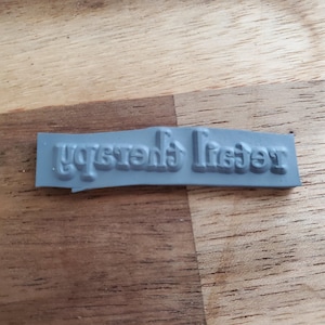 Puede incluir: Un sello de goma gris con el texto "Brenda's Letter" en una fuente serif. El sello es rectangular y el texto está en relieve, en 3D. El sello está sobre una superficie de madera.