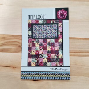 Op de afbeelding: Een quiltpatroon met de titel "Heirloom" van Villa Rosa Designs. Het patroon heeft een patchwork-ontwerp met een verscheidenheid aan bloemenstoffen in tinten rood, roze, geel en blauw.