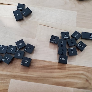 Pode incluir: Uma coleção de teclas de teclado de computador pretas com vários números, letras e símbolos impressos nelas. As teclas estão dispostas em um padrão aleatório sobre uma superfície de madeira marrom claro.