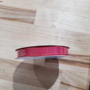 Könnte beinhalten: Eine Spule mit rotem Band. Das Band hat eine einfarbige, leuchtend rote Farbe und scheint aus einem gewebten Material zu bestehen. Die Spule ist rund und hat eine transparente Kunststoffabdeckung. Das Band wird wahrscheinlich für Bastelarbeiten, Geschenkverpackungen oder Dekorationen verwendet.