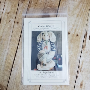 Könnte beinhalten: Ein Nähmuster für ein Stofftier-Kaninchen. Das Muster ist für ein 45 cm großes Kaninchen mit beweglichen Ohren, Armen und Beinen. Das Kaninchen trägt ein blau-weiß gestreiftes Outfit und hat einen beschwerten Boden. Das Muster trägt den Titel "Jr. Rag Rabbit" und wird von "Cotton Ginny's" präsentiert.