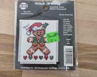 May include: A cross-stitch kit for a gingerbread boy ornament. The kit includes Aida fabric, yarn, and instructions. The gingerbread boy is wearing a Santa hat and a green vest. The text "How Many?" is written on the fabric.