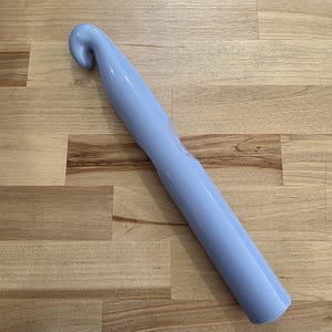 Op de afbeelding: Een grote, lichtblauwe haaknaald met een gebogen kop en een lange, cilindrische steel. De haaknaald is gemaakt van een glad materiaal en is ontworpen om mee te haken. Hij ligt op een houten oppervlak.