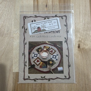 Puede incluir: Auntie Ju's Quilt Shoppe #184 Quilt Block Candle Mat. El paquete muestra un tapete de vela circular con un portavelas central rodeado de bloques acolchados en varios colores y patrones. El tapete tiene aproximadamente 20 cm de diámetro.