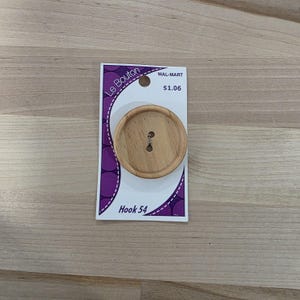 May include: A large, round wooden button with two holes, attached to a white and purple card. The card has the text "Le Bouton" and "Hook 54". The button is light brown and has a natural wood grain.