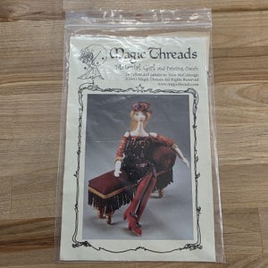May include: A clear plastic bag containing a pattern for a 24-inch cloth doll. The pattern features a doll in a burgundy dress and red shoes, seated on a matching fainting couch. The text reads "Magic Threads Material Girl and Fainting Couch".