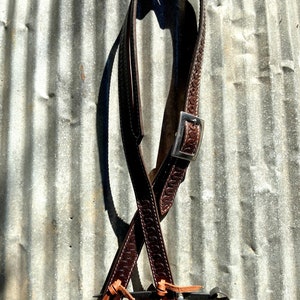 May include: A brown leather strap with a silver buckle and decorative stitching. The strap is hanging on a corrugated metal surface.