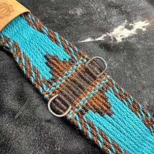 May include: Close-up of a turquoise and brown woven cinch strap with a geometric pattern. The strap features a leather piece with stamped text and two silver metal D-rings. The design is set against a dark, textured background.