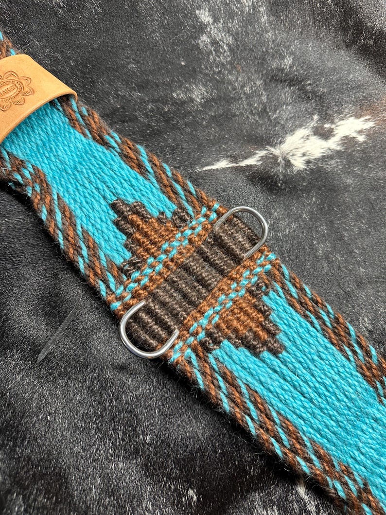 May include: Close-up of a turquoise and brown woven cinch strap with a geometric pattern. The strap features a leather piece with stamped text and two silver metal D-rings. The design is set against a dark, textured background.