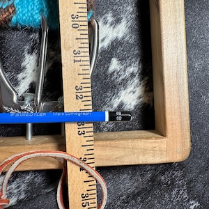 May include: Close-up of a silver-colored belt buckle with a turquoise and brown woven belt. A wooden ruler shows measurements in inches, and a blue pencil is placed across the buckle. A leather strap is also visible.
