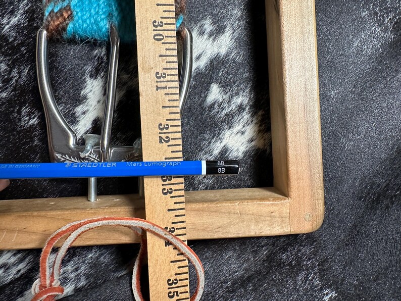 May include: Close-up of a silver-colored belt buckle with a turquoise and brown woven belt. A wooden ruler shows measurements in inches, and a blue pencil is placed across the buckle. A leather strap is also visible.