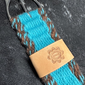 May include: A turquoise and brown woven cinch strap with a silver buckle and a light brown leather accent. The leather accent is stamped with the text "LISA FLOREY MAKER".