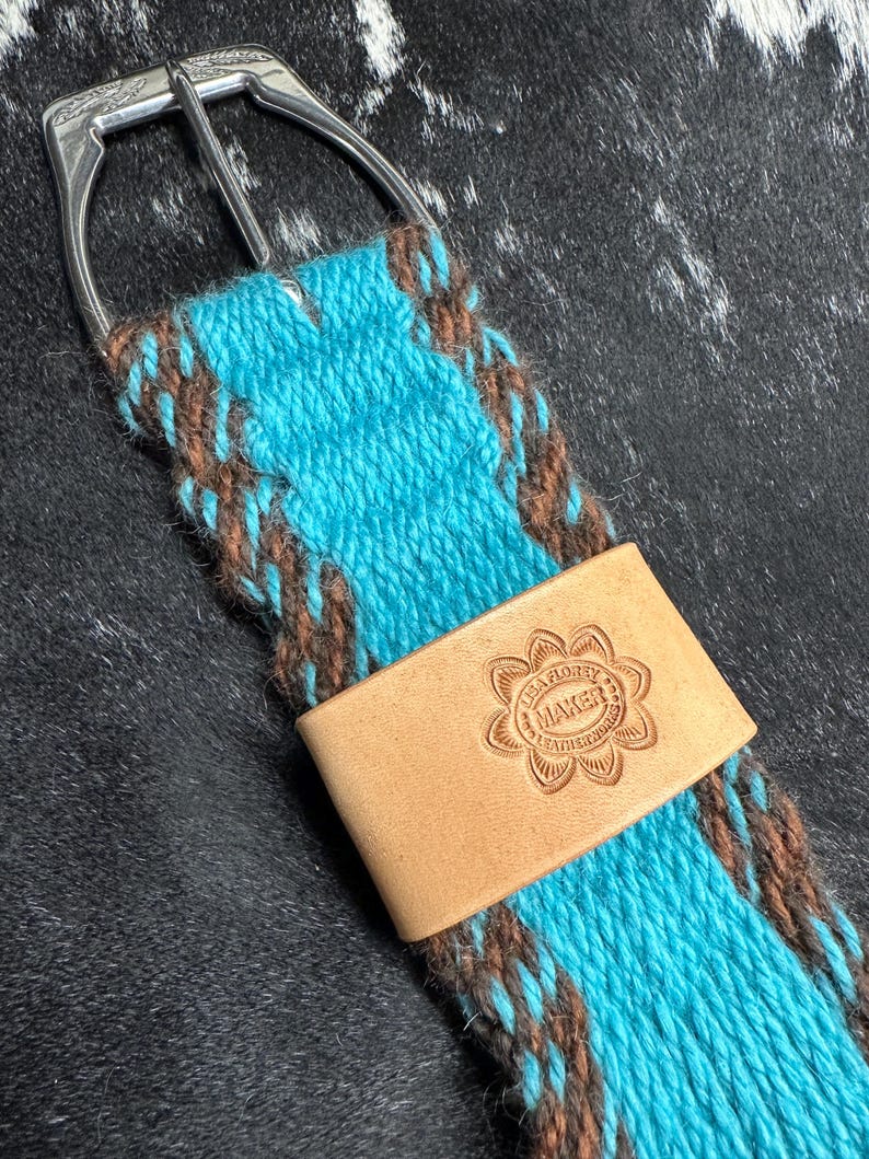 May include: A turquoise and brown woven cinch strap with a silver buckle and a light brown leather accent. The leather accent is stamped with the text "LISA FLOREY MAKER".