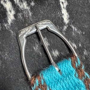 May include: Close-up of a silver-colored belt buckle with a turquoise and brown woven strap. The buckle has a decorative leaf design. The strap is made of a woven material.