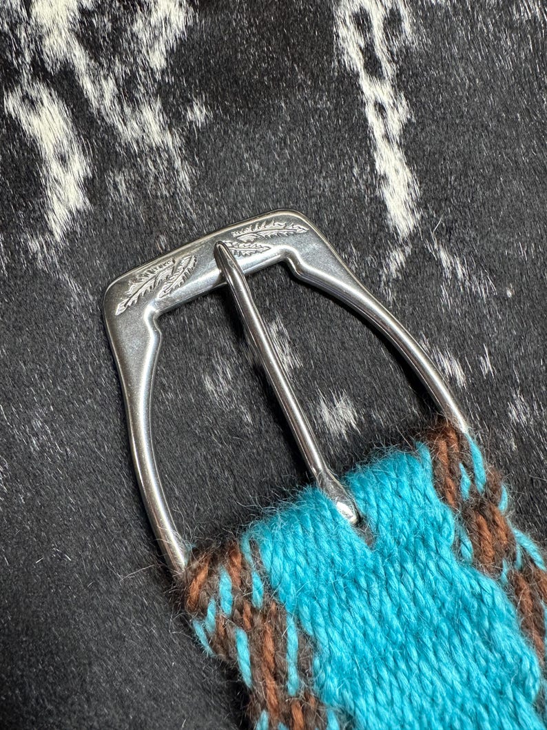 May include: Close-up of a silver-colored belt buckle with a turquoise and brown woven strap. The buckle has a decorative leaf design. The strap is made of a woven material.