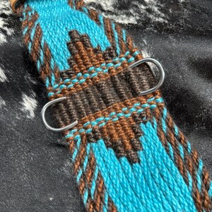 May include: A close-up of a turquoise and brown woven horse cinch. The cinch features a geometric pattern with silver D-rings and a leather strap. The background is a black and white cowhide pattern.
