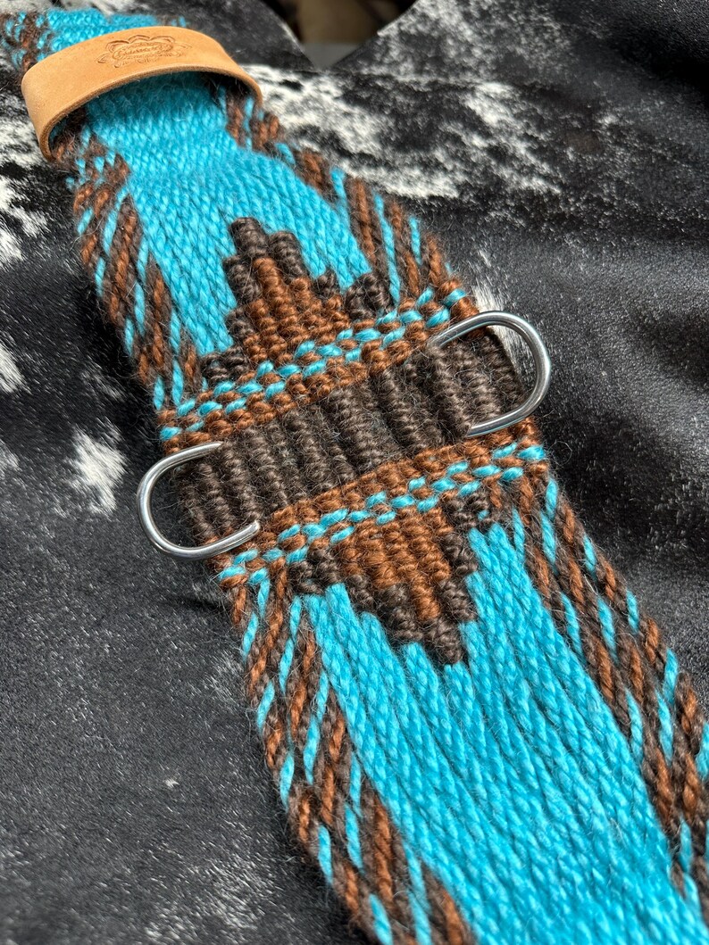 May include: A close-up of a turquoise and brown woven horse cinch. The cinch features a geometric pattern with silver D-rings and a leather strap. The background is a black and white cowhide pattern.